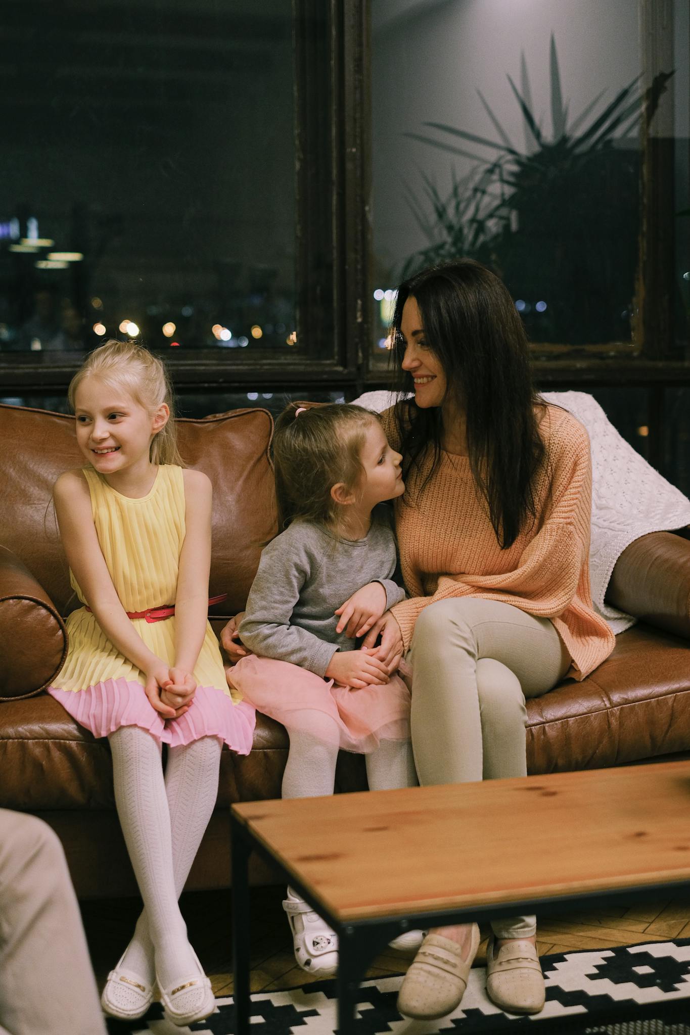 A happy family moment with a mother and her two daughters sitting together indoors on a sofa, sharing smiles and joy.