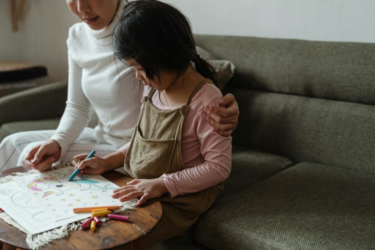 Side view of concentrated little Asian girl in casual outfit sitting at coffee table together with mom while coloring picture with colorful wax crayons